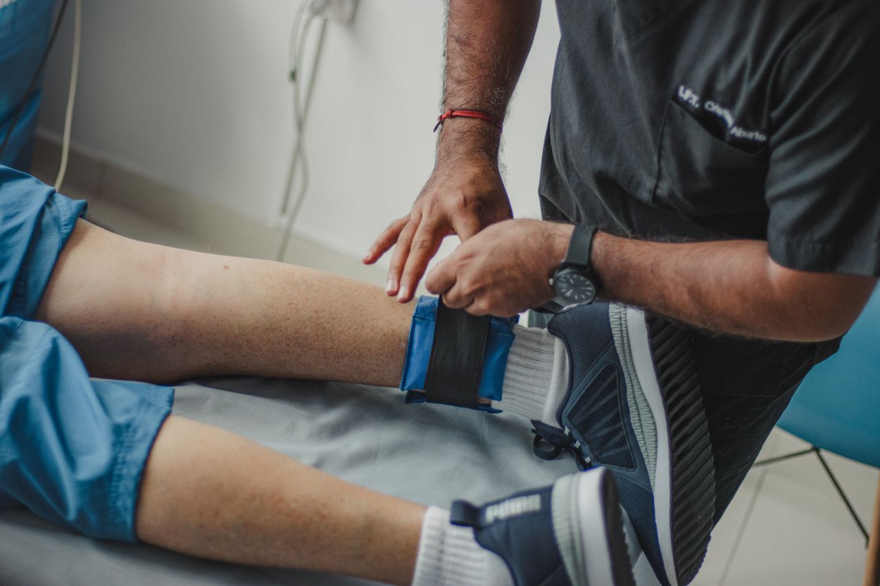 hero-img-01 A physiotherapist adjusts a leg strap on a patient in a clinical setting.